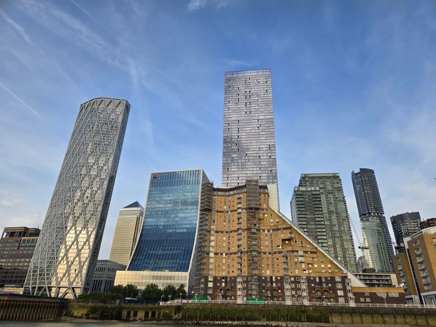 A wide view of the Canary Wharf skyline in London, featuring tall modern office buildings with glass facades and contemporary architectural designs. The buildings are situated along a waterway, with r