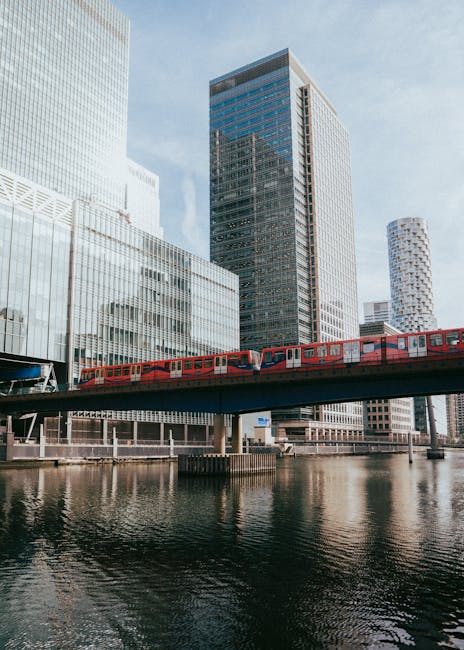 A modern red train is seen moving across a bridge over a river in Canary Wharf, surrounded by tall office buildings with glass facades reflecting the sky. The train appears to be a part of the local t