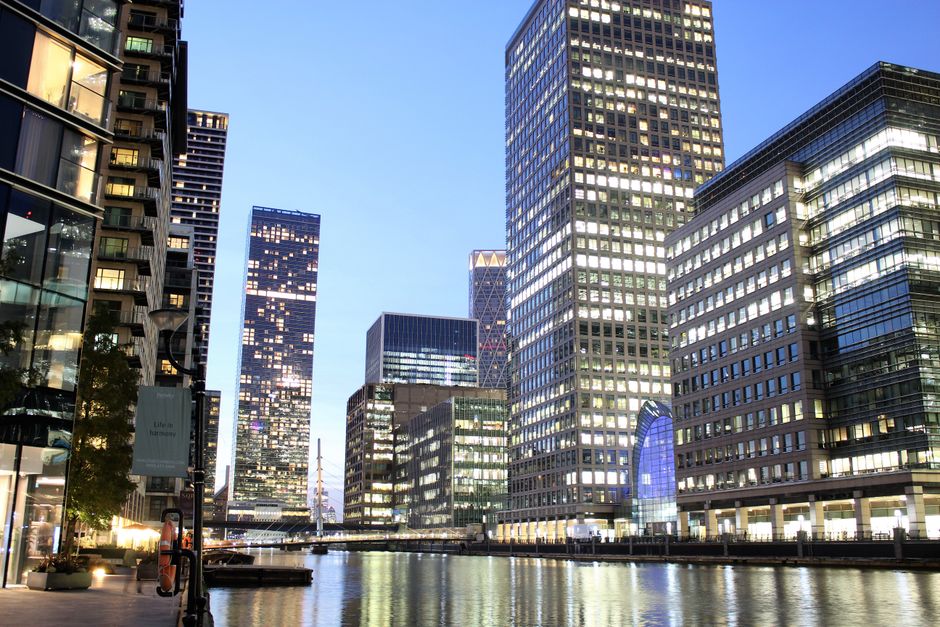 A modern red train is seen moving across a bridge over a river in Canary Wharf, surrounded by tall office buildings with glass facades reflecting the sky. The train appears to be a part of the local t