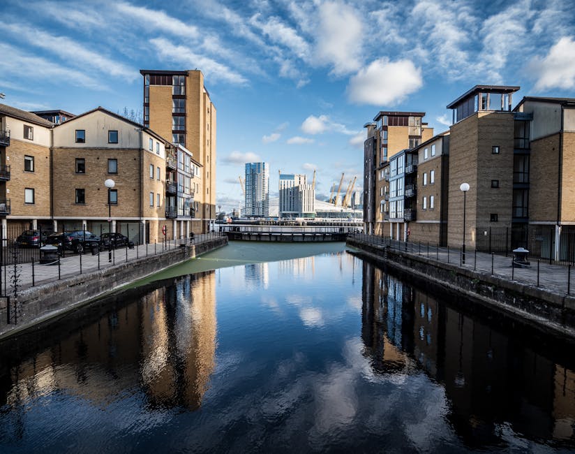 A clear view of the London skyline featuring modern skyscrapers and office buildings under a partly cloudy sky. In the foreground, there is a dock area along the river with blue metal railings, safety